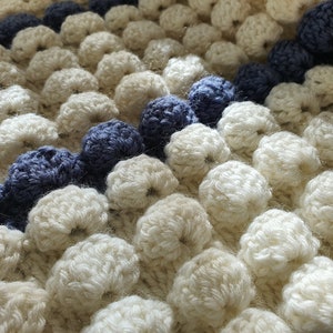 May include: Close-up of a white crocheted blanket with a blue and white striped border. The blanket is made with a textured stitch that creates a bumpy surface.