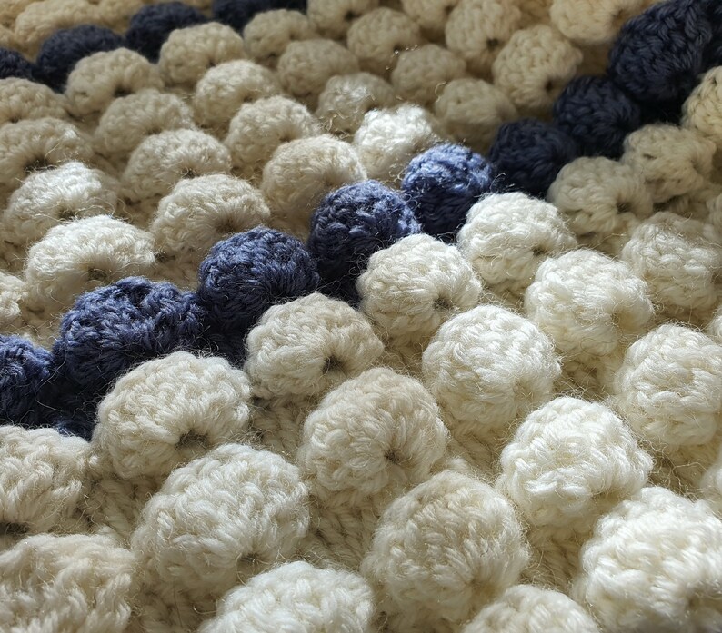 May include: Close-up of a white crocheted blanket with a blue and white striped border. The blanket is made with a textured stitch that creates a bumpy surface.