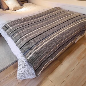 A gray and white striped knitted throw blanket with black and blue stripes. The blanket is folded and placed on a bed.
