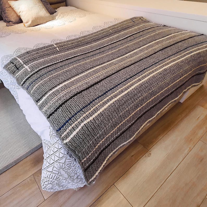 A gray and white striped knitted throw blanket with black and blue stripes. The blanket is folded and placed on a bed.