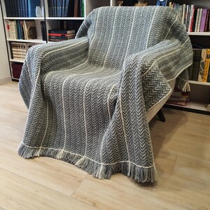 A gray and white crochet striped throw blanket with fringe on the edges. The blanket is draped over an armchair.