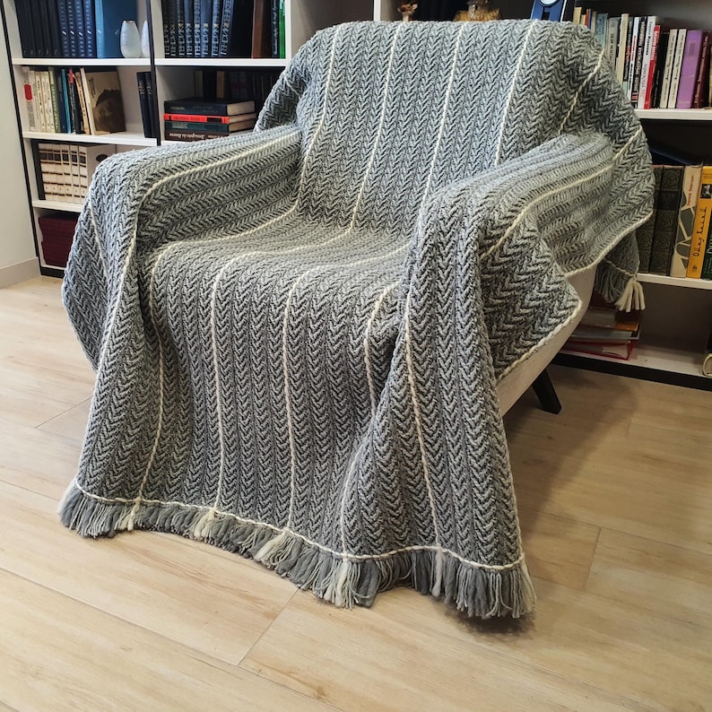 A gray and white crochet striped throw blanket with fringe on the edges. The blanket is draped over an armchair.