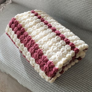 A white and burgundy crocheted blanket with a textured, puffy design. The blanket is folded in half and has a striped pattern.