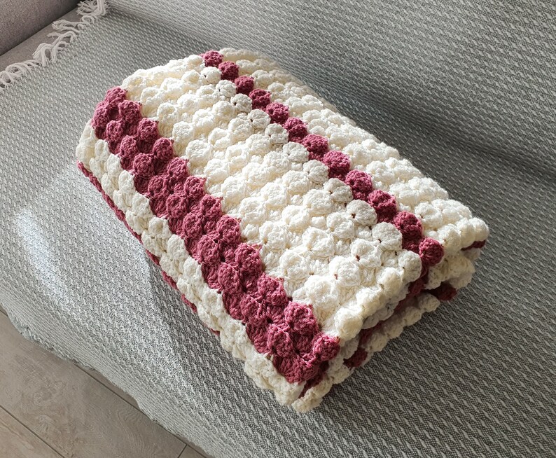A white and burgundy crocheted blanket with a textured, puffy design. The blanket is folded in half and has a striped pattern.