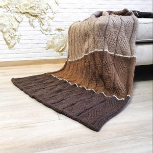 A brown, beige, and white knitted throw blanket with a cable knit pattern. The blanket is draped over a couch.