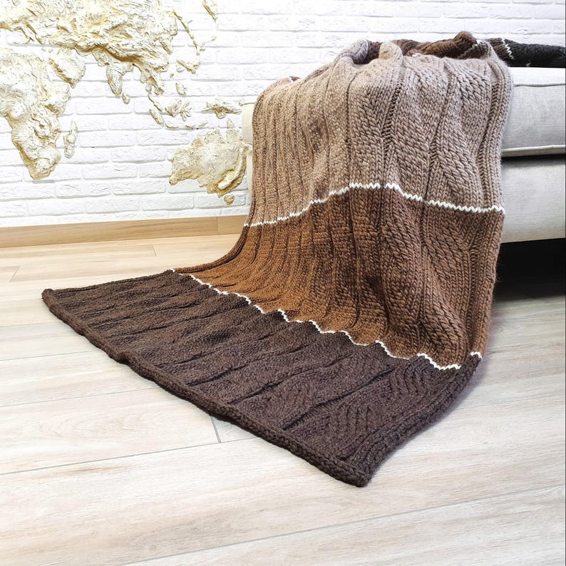 A brown, beige, and white knitted throw blanket with a cable knit pattern. The blanket is draped over a couch.