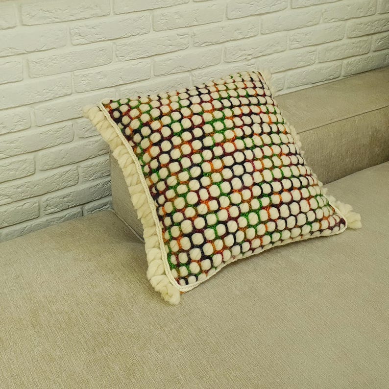K&ouml;nnte beinhalten: Ein quadratisches Zierkissen mit wei&szlig;em Hintergrund und einem Muster aus bunten Punkten in Gr&uuml;n, Orange und Lila. Das Kissen hat einen cremefarbenen R&uuml;schenrand und liegt auf einem beigen Sofa.