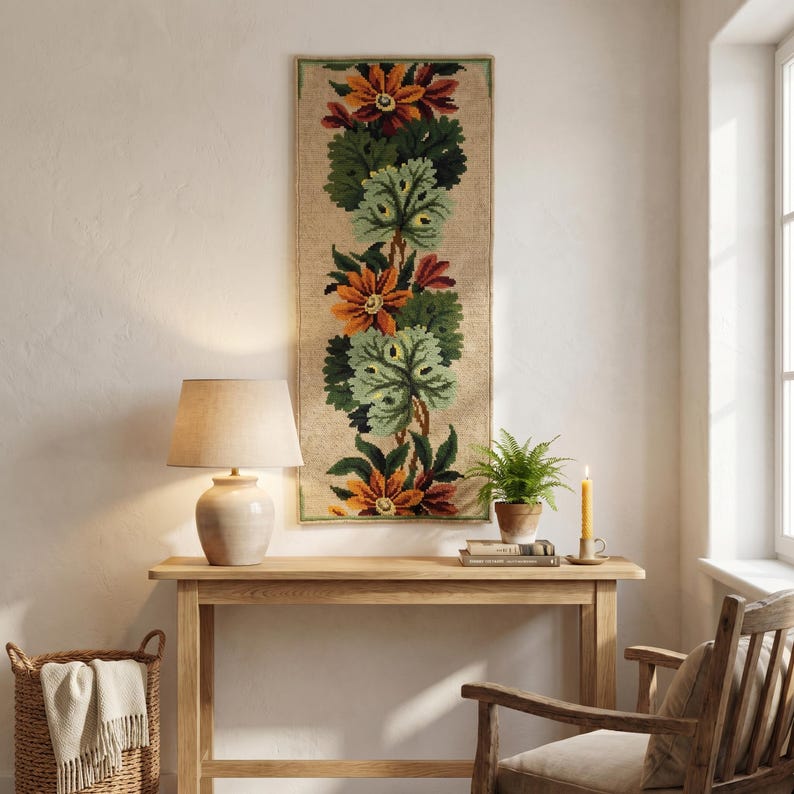 Handmade wool embroidery panel featuring floral pattern with green leaves, decorative wall hanging on the light wall. over a small table.