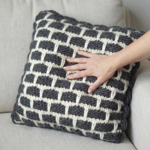 May include: A square cushion featuring a grey and cream geometric design. The cushion is made from a thick, textured fabric and is placed on a light-coloured sofa. The pattern is a repeating grid-like design.