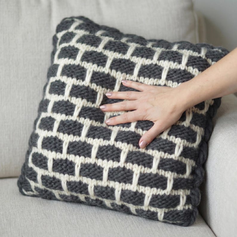 May include: A square cushion featuring a grey and cream geometric design. The cushion is made from a thick, textured fabric and is placed on a light-coloured sofa. The pattern is a repeating grid-like design.