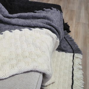 A white, gray, and black knitted throw blanket with fringe on the edges. The blanket is folded and draped over a couch.