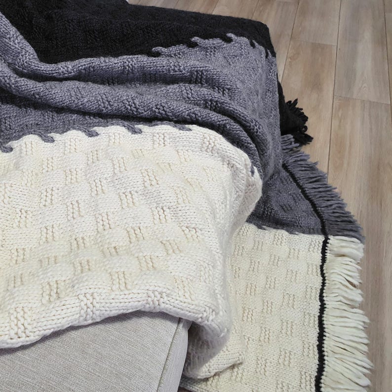 A white, gray, and black knitted throw blanket with fringe on the edges. The blanket is folded and draped over a couch.