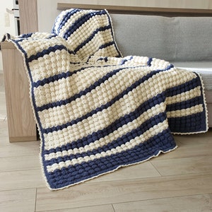 May include: A white and blue striped crocheted throw blanket with a textured bobble stitch. The blanket is draped over a couch.