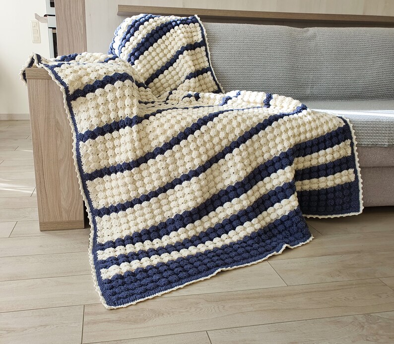 May include: A white and blue striped crocheted throw blanket with a textured bobble stitch. The blanket is draped over a couch.