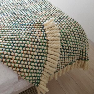 A textured, cream-colored blanket with a green dotted pattern and cream-colored fringe is draped on a bed showing its becking. The blanket is made of a soft material and adds a cozy touch to the room. The blanket is approximately 60 inches long.