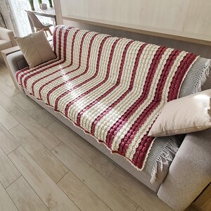 A white and pink striped crocheted throw blanket with fringe on a gray couch. The blanket has a textured, bumpy design.