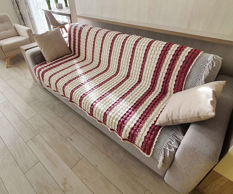 A white and pink striped crocheted throw blanket with fringe on a gray couch. The blanket has a textured, bumpy design.