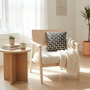 May include: A light wood armchair with a cream cushion and a decorative pillow with a black and white brick pattern. A cream throw blanket is draped over the arm. A small round side table holds a potted plant, books, and a mug.