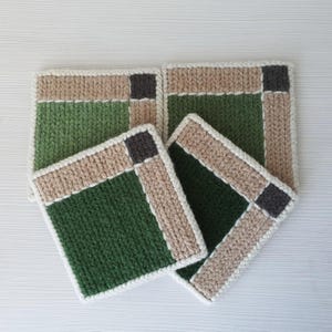 May include: Four square, hand-knitted coasters. Each coaster features a green center, bordered by a tan and white knitted pattern. A small, dark gray square accents each corner. These coasters are perfect for protecting surfaces from hot or cold beverages.