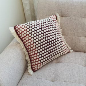 May include: Square decorative pillow with a white bobble pattern over a burgundy background. The pillow has a fringe trim and is resting on a beige sofa. The pillow is a home decor item.