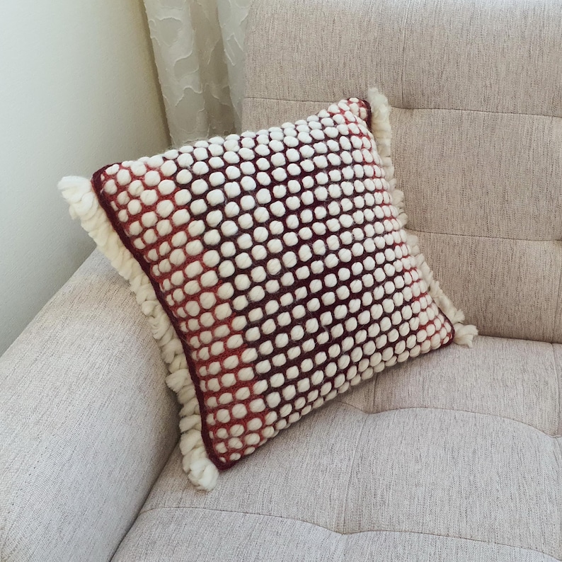May include: Square decorative pillow with a white bobble pattern over a burgundy background. The pillow has a fringe trim and is resting on a beige sofa. The pillow is a home decor item.