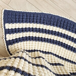 May include: A crocheted blanket with a white and blue striped pattern. The blanket is made with a textured stitch that creates a bumpy surface.