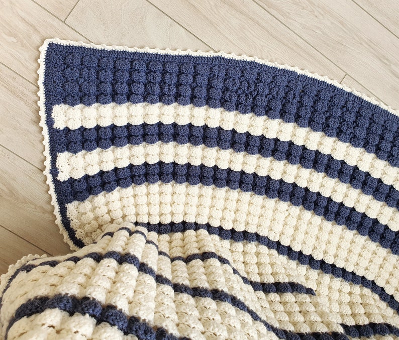 May include: A crocheted blanket with a white and blue striped pattern. The blanket is made with a textured stitch that creates a bumpy surface.