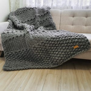 A chunky knit gray blanket draped over a beige sofa. The blanket features a textured pattern and a small wooden tag. The thick yarn creates a cozy, inviting look, perfect for home decor and comfort.