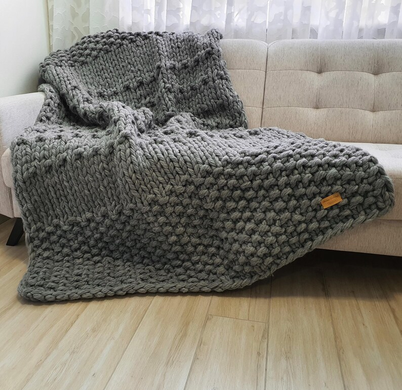A chunky knit gray blanket draped over a beige sofa. The blanket features a textured pattern and a small wooden tag. The thick yarn creates a cozy, inviting look, perfect for home decor and comfort.