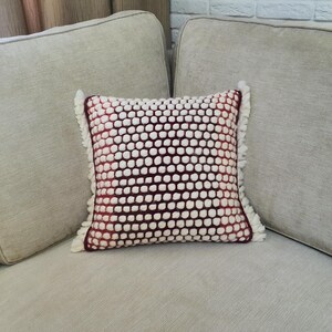 May include: Square decorative pillow with a textured design. The pillow features a pattern of white bobbles on a burgundy background. The edges are trimmed with white fringe. The pillow is placed on a light gray sofa.