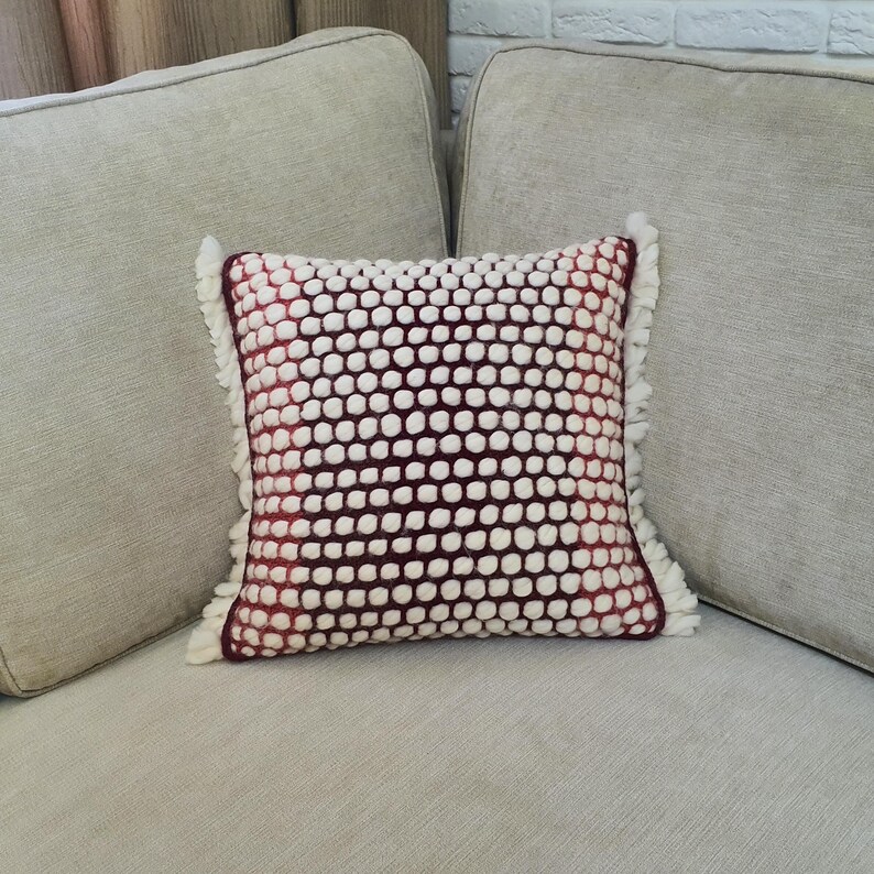 May include: Square decorative pillow with a textured design. The pillow features a pattern of white bobbles on a burgundy background. The edges are trimmed with white fringe. The pillow is placed on a light gray sofa.