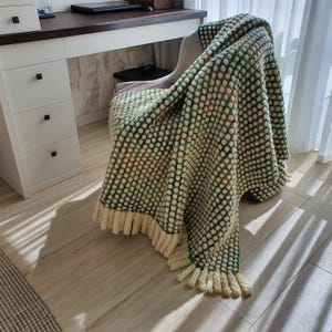 A textured, cream-colored blanket with a green dotted pattern and cream-colored fringe is draped over a chair. The blanket is made of a soft material and adds a cozy touch to the room. The blanket is approximately 60 inches long.