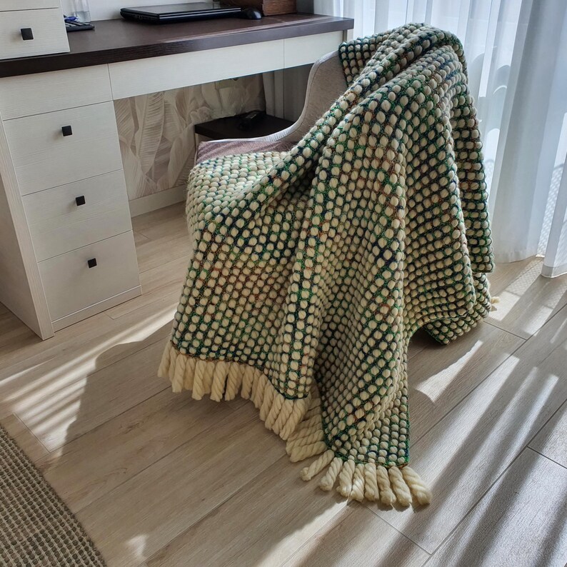 A textured, cream-colored blanket with a green dotted pattern and cream-colored fringe is draped over a chair. The blanket is made of a soft material and adds a cozy touch to the room. The blanket is approximately 60 inches long.