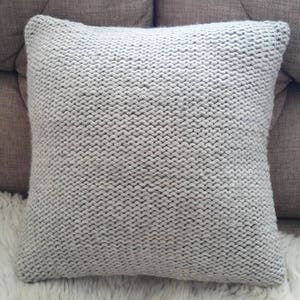 May include: A square, light grey knitted cushion. The cushion has a chunky knit texture and is placed on a white, fluffy surface. The cushion is a neutral colour and would complement various home decor styles.