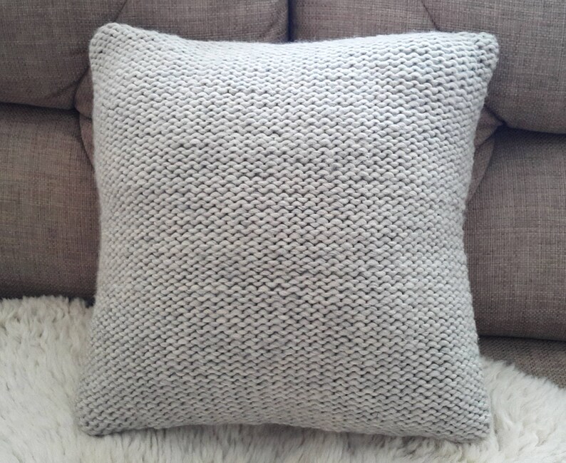 May include: A square, light grey knitted cushion. The cushion has a chunky knit texture and is placed on a white, fluffy surface. The cushion is a neutral colour and would complement various home decor styles.