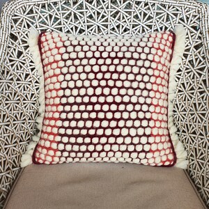 May include: Square decorative pillow with a white and burgundy dotted pattern. The pillow has a fringe trim on the sides. The pillow is on a woven chair.