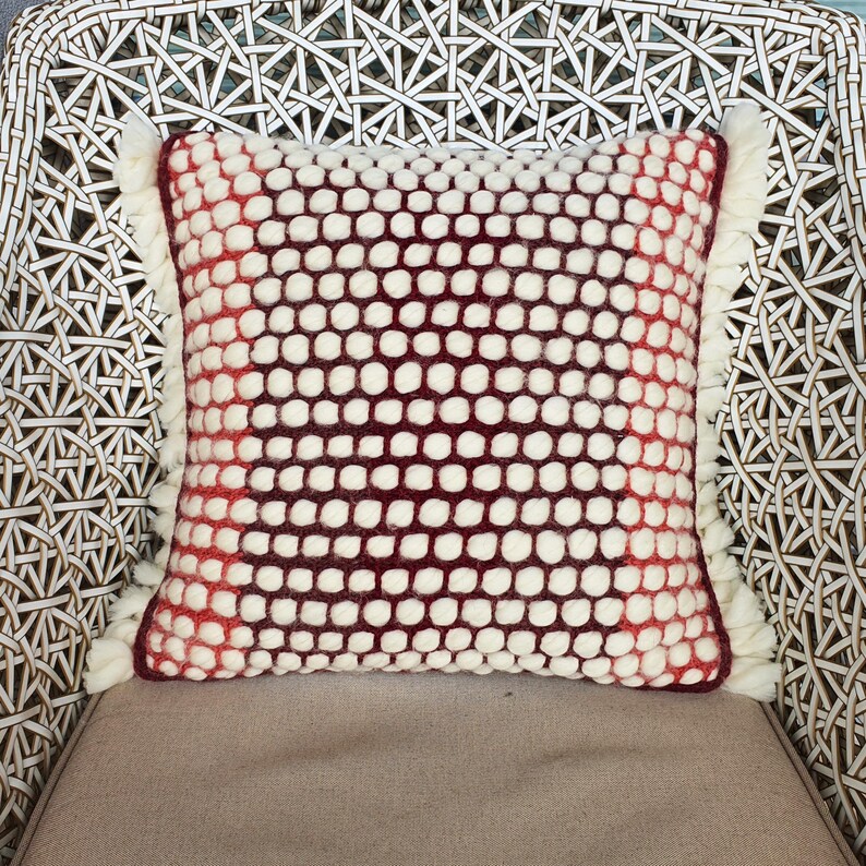 May include: Square decorative pillow with a white and burgundy dotted pattern. The pillow has a fringe trim on the sides. The pillow is on a woven chair.