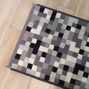 K&ouml;nnte beinhalten: Ein geometrischer Teppich in Grau, Schwarz und Wei&szlig; mit einem grauen Rand. Der Teppich h&auml;ngt an einer Metallstange.