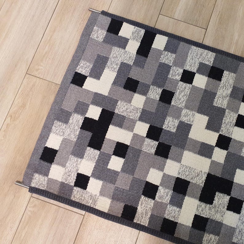 K&ouml;nnte beinhalten: Ein geometrischer Teppich in Grau, Schwarz und Wei&szlig; mit einem grauen Rand. Der Teppich h&auml;ngt an einer Metallstange.