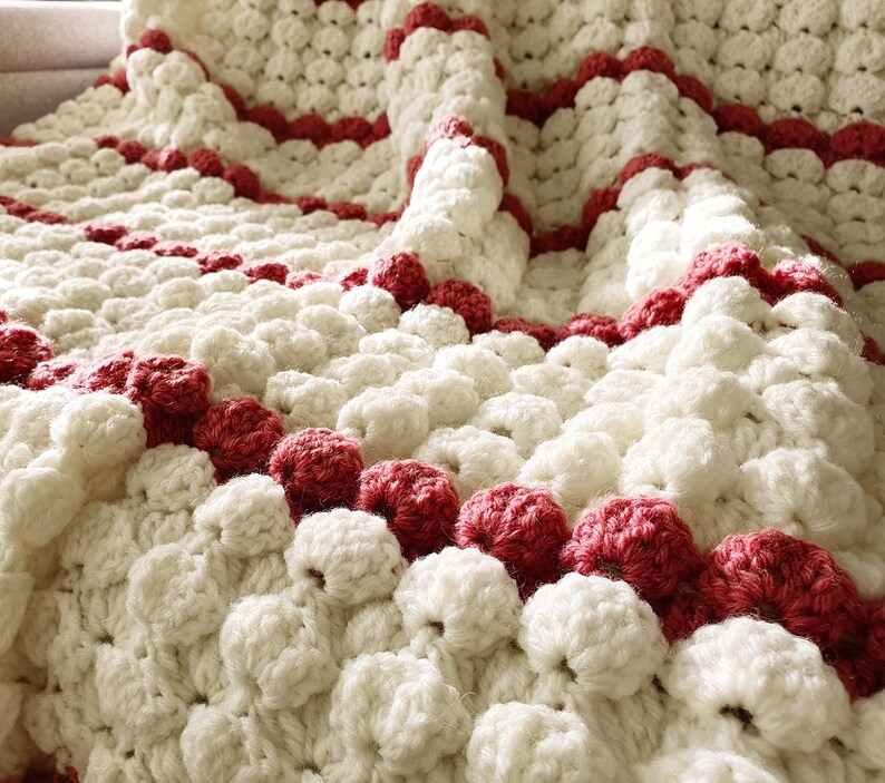 Close-up a white and red crocheted blanket with a textured, puffy design. The blanket has a repeating pattern of white circles with red stripes.