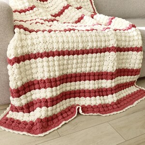 A crocheted throw blanket with a striped pattern in white and burgundy. The blanket has a textured, puffy surface and a scalloped edge.