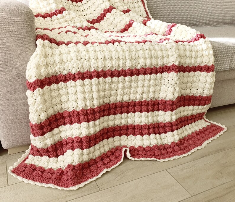 A crocheted throw blanket with a striped pattern in white and burgundy. The blanket has a textured, puffy surface and a scalloped edge.