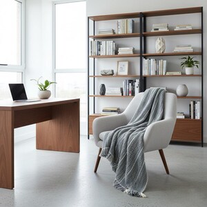 May include: A modern office space featuring a wooden desk with a laptop and a potted plant. A gray armchair with a patterned throw blanket sits near a large bookshelf filled with books and decorative items. The room has a minimalist design.