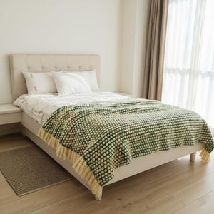 A bed with a beige headboard and white bedding. A green and cream-colored textured throw blanket with fringe is draped over the bed. A small rug sits on the wooden floor.