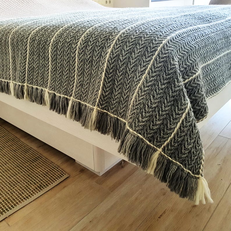 May include: A gray and white herringbone pattern throw blanket with fringe. The blanket is draped over a bed, showcasing its texture and design. The fringe is a mix of gray and white, adding a decorative element to the blanket.