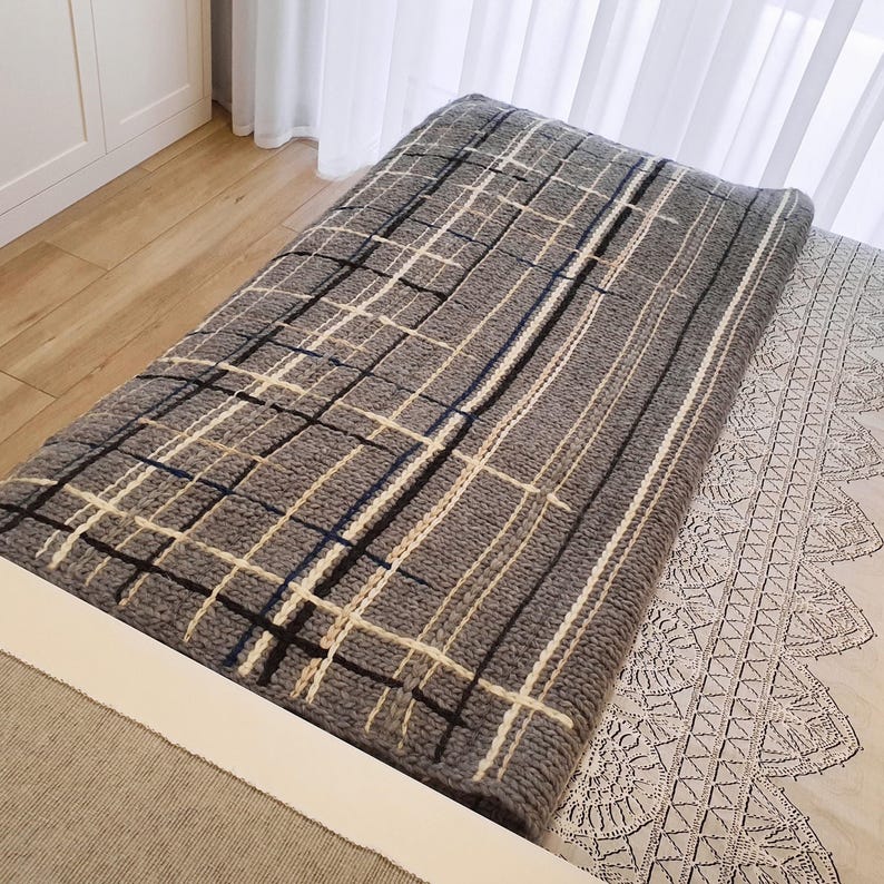 A gray and white patterned throw blanket with a black and white geometric design. The blanket is folded and placed on a white bed with a white lace trim.