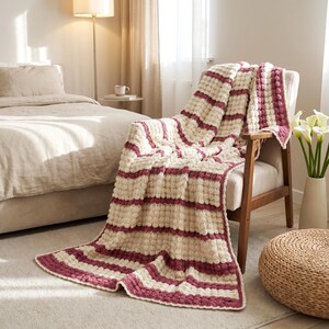 A crocheted throw blanket with white and burgundy stripes. The blanket is draped over a light brown armchair in a cozy bedroom.