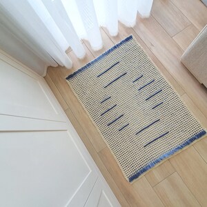 May include: A cream-colored crocheted rug with a blue geometric pattern. The rug has a fringe border and is laid out on a wooden floor.