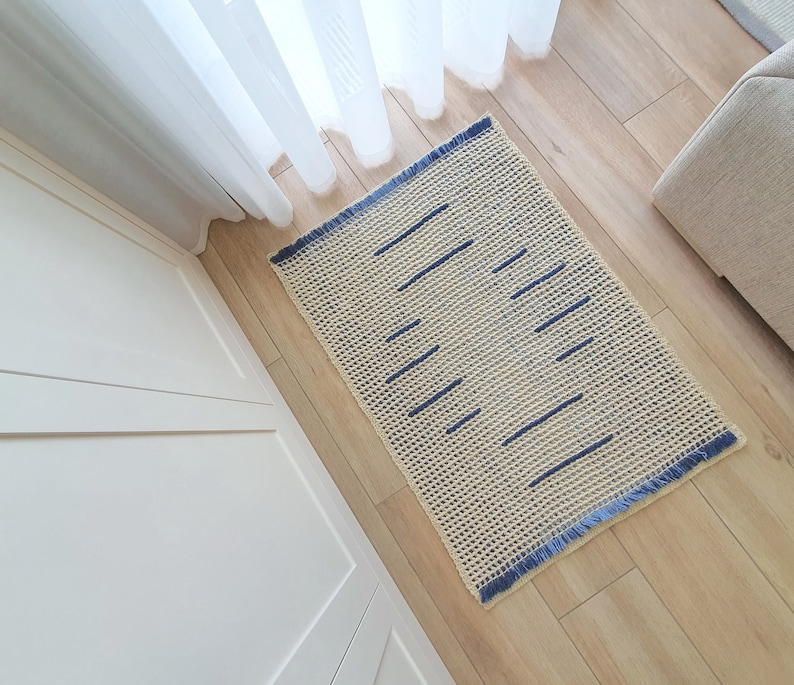 May include: A cream-colored crocheted rug with a blue geometric pattern. The rug has a fringe border and is laid out on a wooden floor.