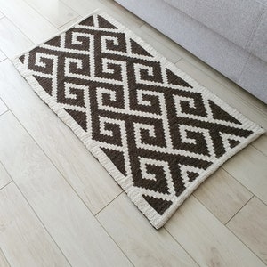 May include: A brown and white geometric patterned rug with a white border. The rug is made of a soft, woven material.